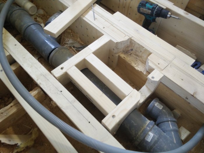 Rakennustyömaa, jossa näkyy puisia tukirakenteita, mustia muoviputkia ja pora, kylpyhuoneen lattian rakentaminen kesken.