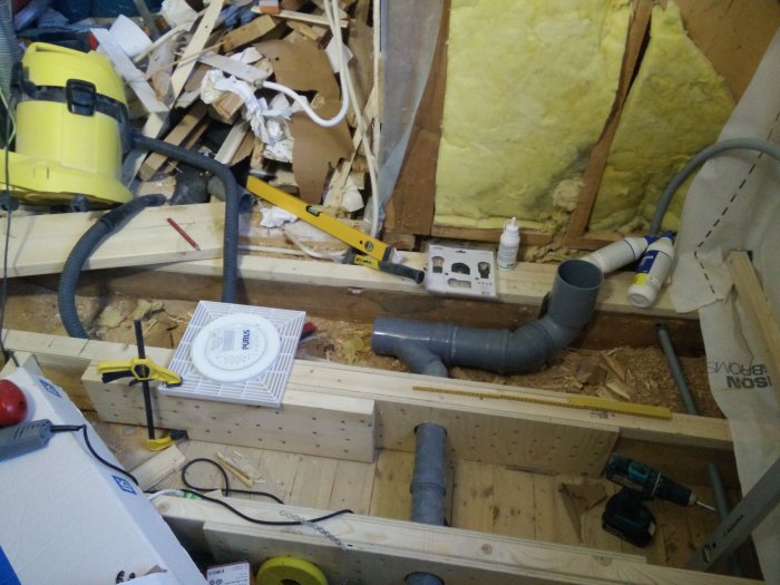 Badrumsrenovering pågår med synliga trästöd, rör och verktyg, inklusive gul dammsugare och borrmaskin. Byggmaterial och isolering i bakgrunden.
