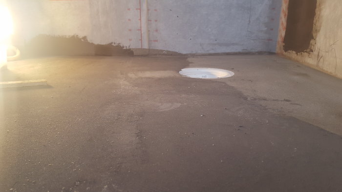 Golv med betongpass nära väggen och golvbrunn som behöver slipas för fall.