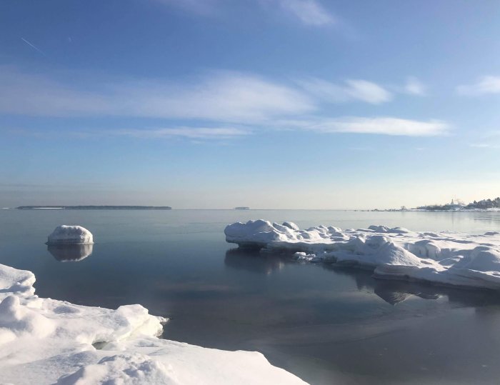 Vinterlandskap med snötäckta isflak på stilla vatten mot en klarblå himmel.