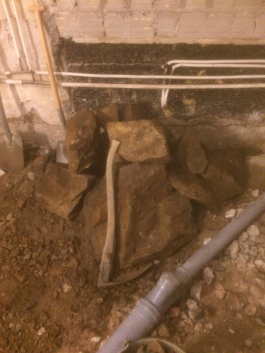 Grovt spett ligger bland större stenar på ett grusigt underlag i ett pågående byggprojekt med synliga rörledningar.