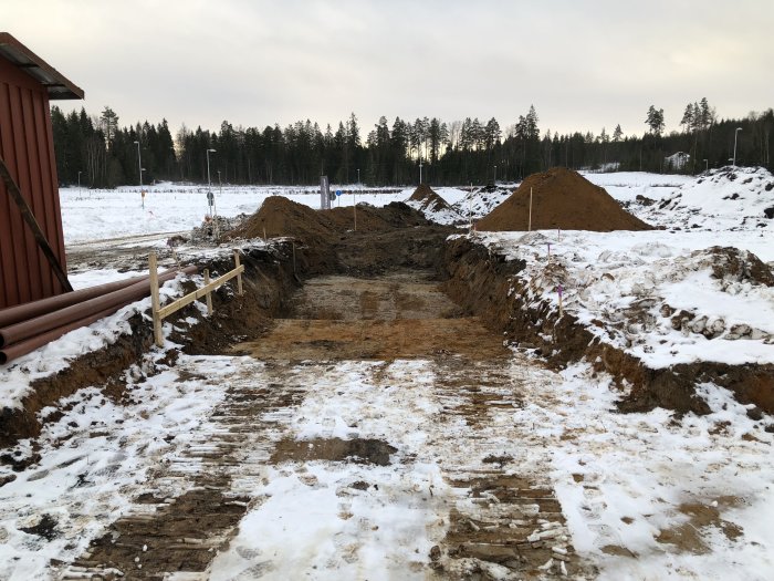 Början på grävningen för husbygge med synlig markarbeten, jordhögar och snötäckt terräng i bakgrunden.