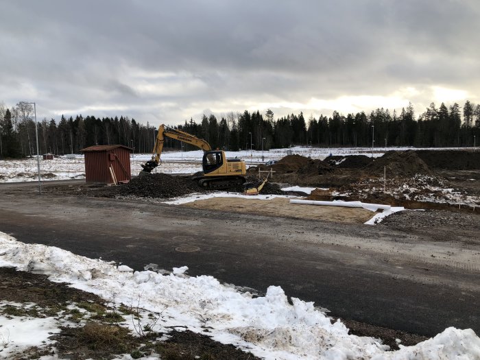 En grävmaskin på en byggplats i början av ett byggprojekt, med högar av grävd jord och snö runtomkring.