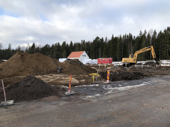 Grävmaskin vid en byggplats med högar av grus och markarbete pågår, omgiven av skog och byggbodar i bakgrunden.