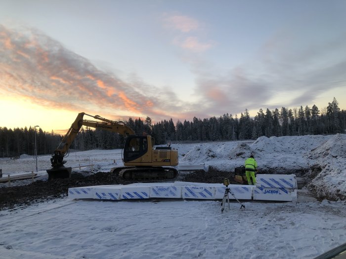 En grävmaskin och en arbetare förbereder marken vid solnedgång för att gjuta en byggplatta.