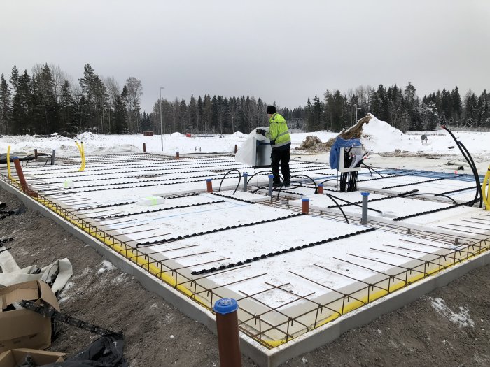 Byggplats med förberedelser för golvgjutning, VVS-installation synlig samt arbetare som mäter.