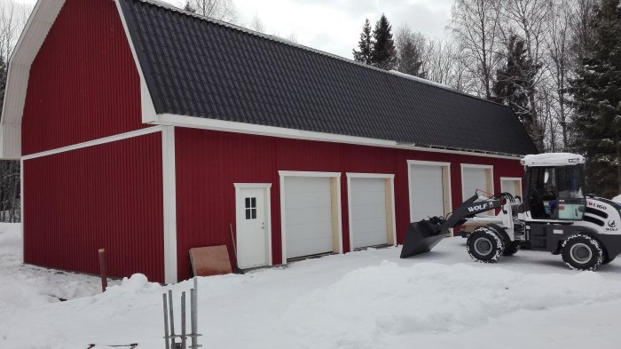 Röd byggnad med svart tak och fyra garageportar, en vit dörr och en hjullastare utanför i snön.