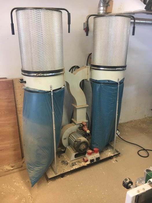 Två industriella dammavskiljare med filter och anslutna blå säckar i ett verkstadsrum fyllt med spån och verktyg.
