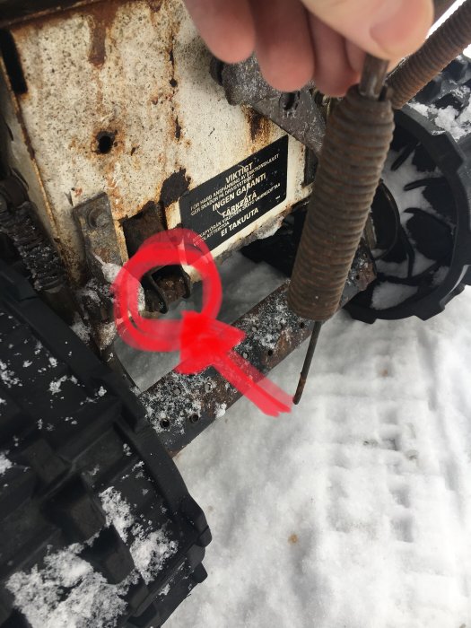 Hand håller avsliten vajer vid maskindel med röd markering på snöig bakgrund.