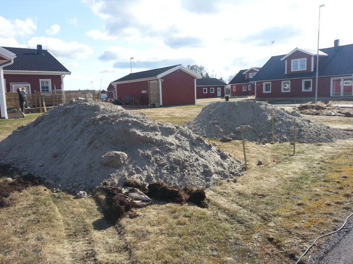 En hög med fyllnadsmassa på en gräsmatta framför röda trähus, med mätstolpar och en delvis synlig person i bakgrunden.