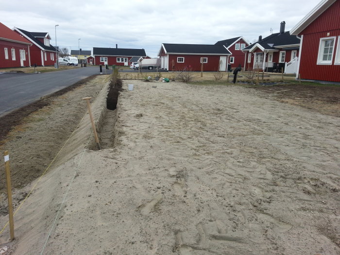 Nyjämnad gräsmattejord framför röda hus med påbörjad plantering längs gatan.