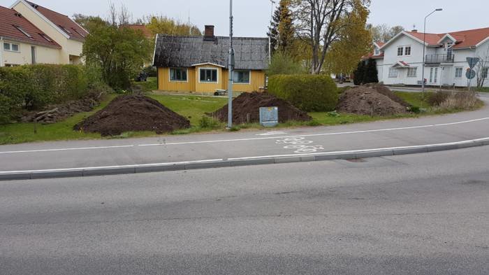 En gul villa med blått tak bakom en väg, omgiven av högar med jord i förgrund, som indikerar landskapsgestaltning.