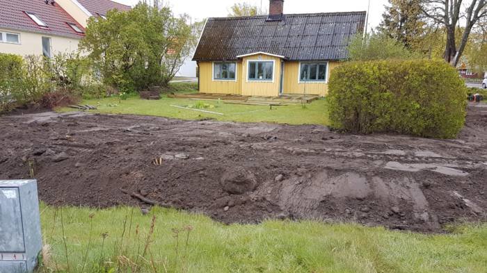 Trädgård under anläggning med ojämn mörk jord framför gult hus, utan gräsmatta och med stenpartier.