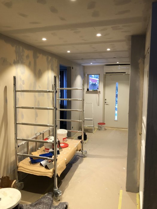 Interiör av ett rum under renovering med spacklade väggar och innertak, ställning och målarutrustning.