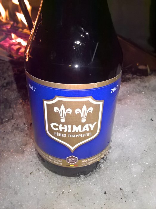 En flaska Chimay-öl i snön framför ett lägereldsglöd.