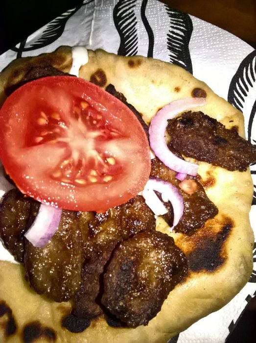 Naanbröd med grillat kebabkött, tomatskiva och lökringar serverat på en tallrik med svartvitt mönster.