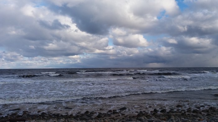 Havsvy med vågor som rullar in på en strand kantad av småsten under en molnig himmel.