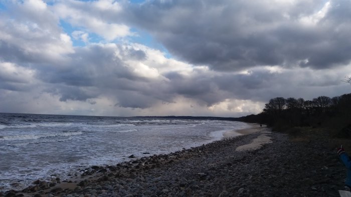 Molnig himmel över en stenig strandlinje med vågor och en person i fjärran.