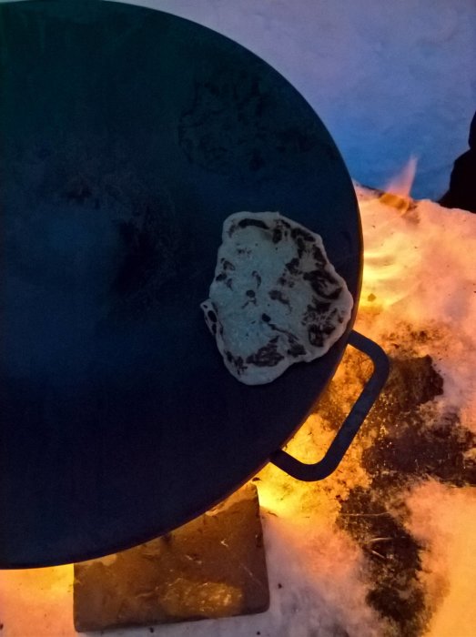 Stekhäll (Muurikka) utomhus i snön med ett naanbröd på, flamman från en eldstad syns i bakgrunden.