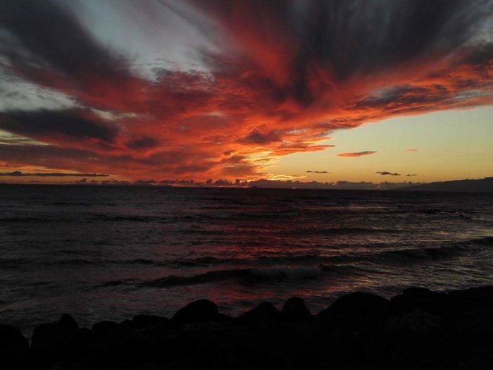 Dramatisk solnedgång med röd-orange himmel över havet, sett från en strand med mörka stenar.