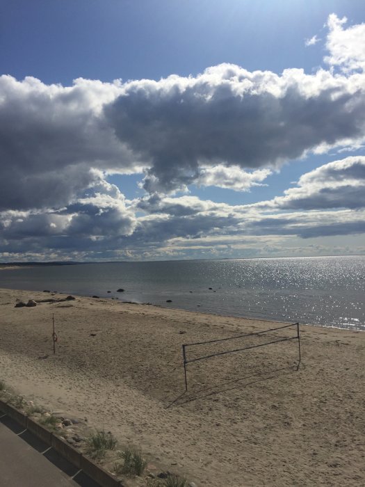 Solbelyst sandstrand med volleybollnät, hav i bakgrunden och en dramatisk himmel med moln.