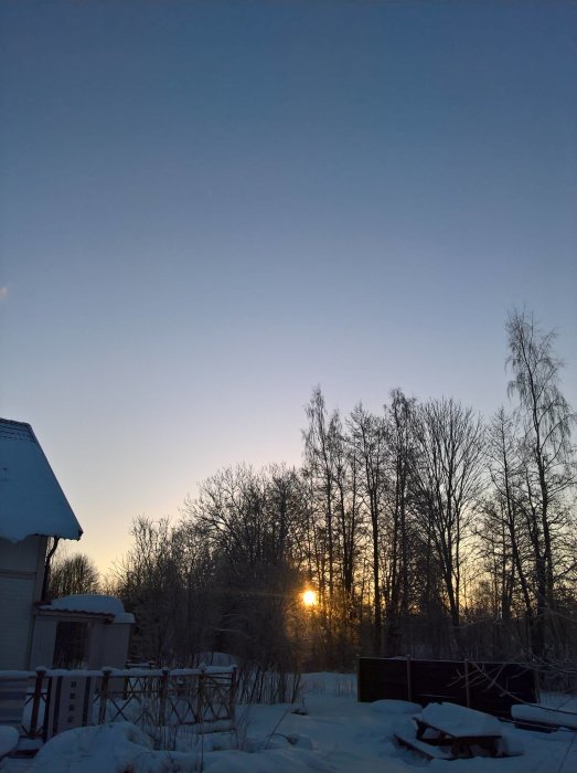 Klar himmel vid soluppgång över ett snötäckt landskap med träd och hus.