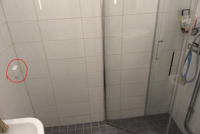 Eluttag markerat med röd cirkel nära duschkabin i badrum med vita kakelväggar och grått klinkergolv.