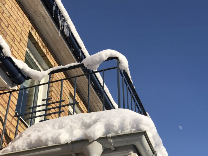 Snötäckt balkongräcke med is istappar mot en klarblå himmel och synlig halvmåne.