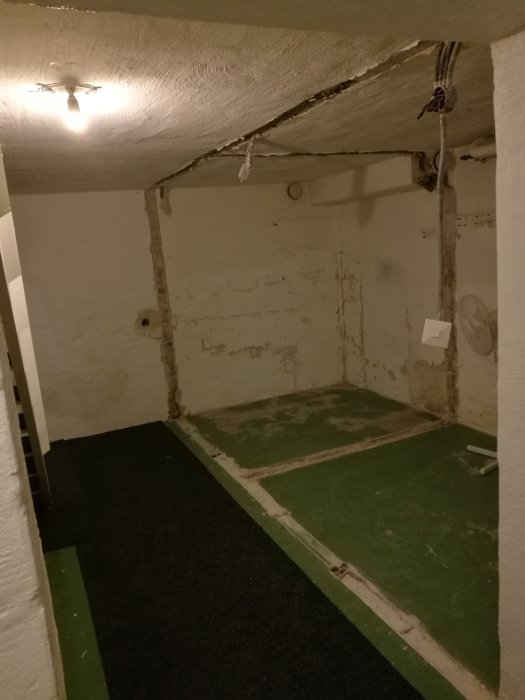 Källare med nyligen rivna väggar som lämnat två rännor i det gröna betonggolvet, intill en svart matta.