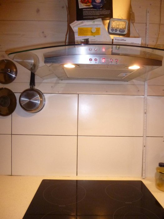 Köksvägg med 50 cm kakel och vitvaxad träpanel ovanför samt en rostfri spisfläkt med magneter och tidtagarur.