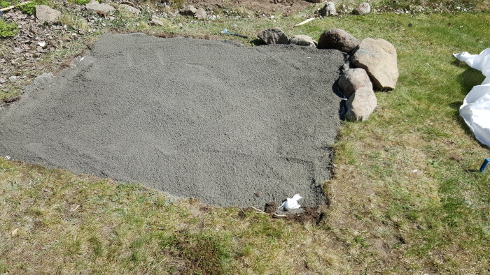 Grund för växthus förbereds med utjämnad fyllnadsmassa på lutande gräsmatta, omgiven av stenar.