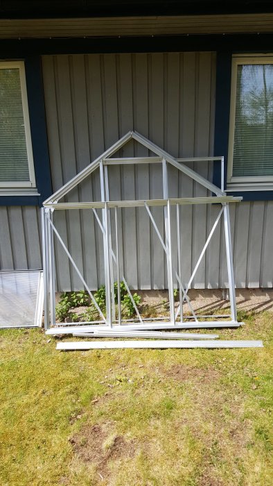 Grundkonstruktion av ett växthus med aluminiumprofiler på ett stenplattsgolv, placerat framför ett hus.