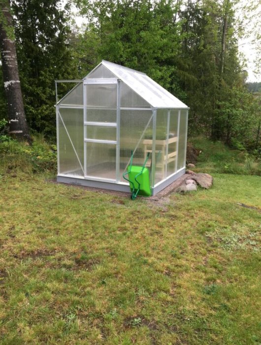 Ett nybyggt växthus med aluminiumprofiler och stenplattsgolv på en lutande backe, med en grön vattenkanna framför.