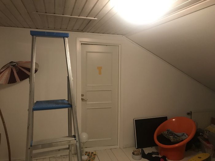 Övervåningsrum med snedtak och vit dörr, blå stege i förgrunden med isolering ovanför, indikerar renovering.