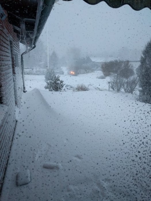 Ett snöigt landskap som täcker ett hus och en trädgård under en snöstorm med svagt synliga konturer av ett fordon.