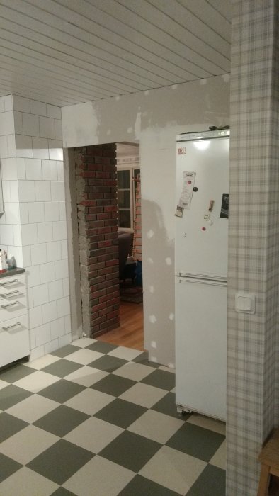Delvis renoverat kök med vit kyl, tegelvägg, vitt kakel och rutigt linoleumgolv.