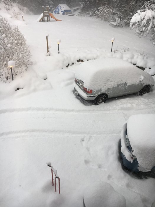 Uppfart täckt av snö med en bil under snötäcke och lekställning i bakgrunden.
