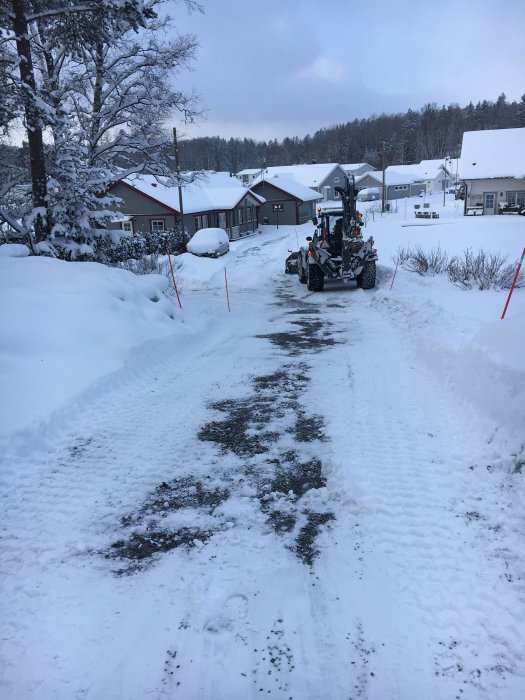 Snöig väg med däckspår och en traktor, hus och träd i bakgrunden.