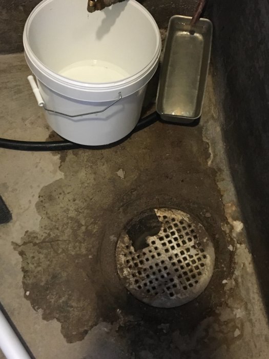 Vattenläckage från kopparrör över hink och golvbrunn på betonggolv med fuktfläckar.