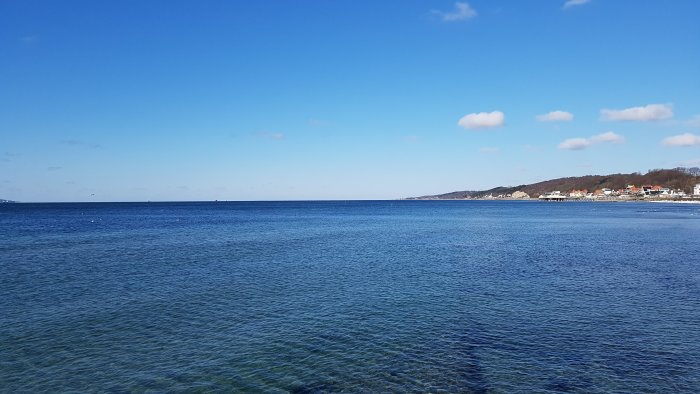 Utsikt över klart havsvatten med strandlinje och hus i bakgrunden under blå himmel.