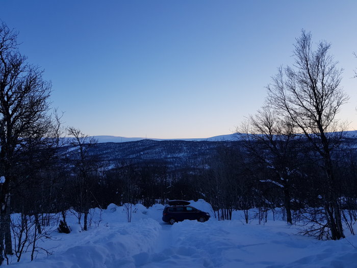 Bil omgiven av djup snö i en skog med berg i bakgrunden vid skymning.