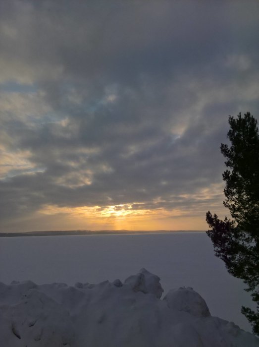 Utsikt från ett fönster med solnedgång över snötäckt landskap och molnhimmel.