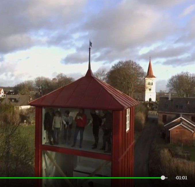 Rödmålat tornliknande utrymme med helglasade väggar och folk inuti, kyrktorn i bakgrunden.