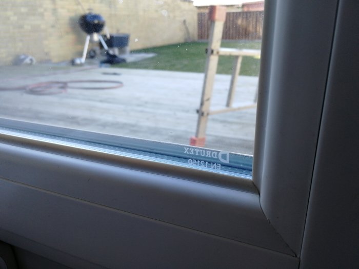 Närbild på en fönsterkarm med texten "DRUTEX" synlig, med utsikt över en trädgård med grill och stegar.