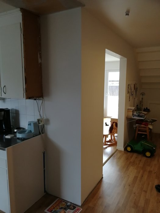 Renoverat kök med delvis spacklad vägg, trägolv och en grön leksakstraktor, skymd hund i bakgrunden.