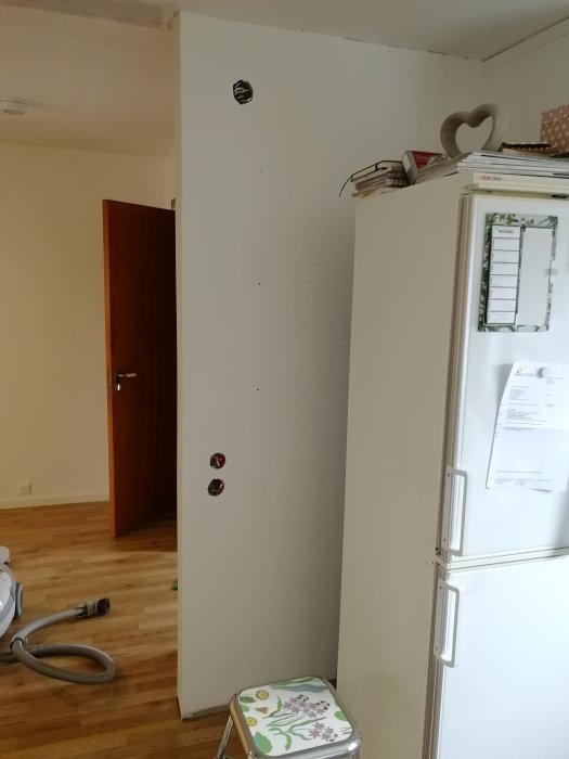Nyrenoverad rumshörn med nymålad vit vägg och orenoverat tak, med en dörr i bakgrunden och kylskåp till höger.