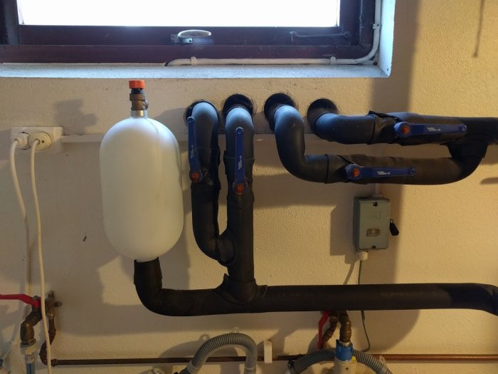 Expansionskärl och rörledningar för jordvärmepump i källare, med ventiler och anslutningar.