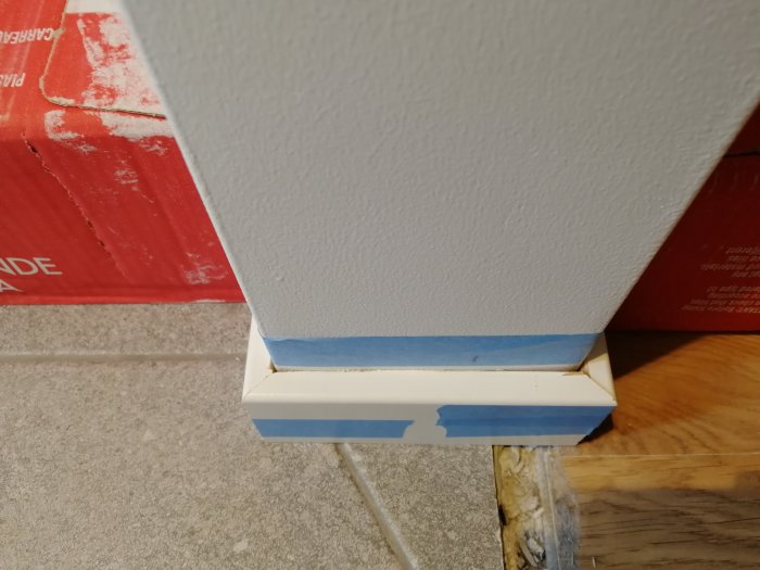Hörn av ett rum med vit listning vid golv, skyddad med blå målartejp, och en klinkerförpackning som tyngd.