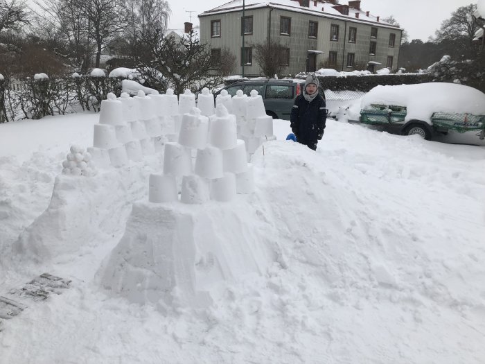 Person står bredvid en snöborg med flera torn och snöblock, på en snötäckt parkering.