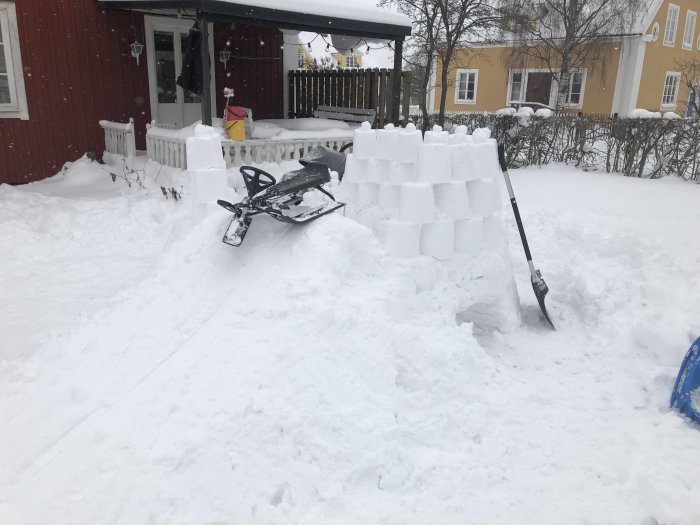 Byggd av snöblock framför röd hus: snöskoter och snöskovlar syns.